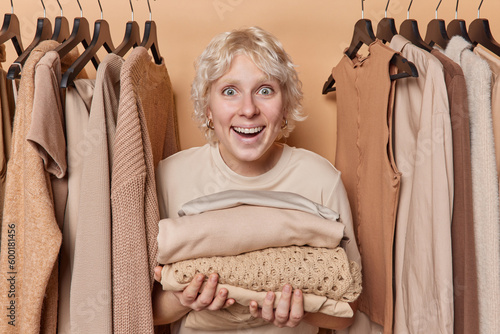 Cheerful surprised young woman holds stack of folded clothes smiles broadly and reacts to something awesome poses among clothig on rails sorts out her wardrobe stands against brown background photo