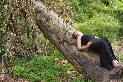Young woman, blonde and beautiful, with a black dress, with her eyes closed, lying on the trunk of a big tree, in the middle of nature. Concept nature, peace, tranquility, trees. © Manuel