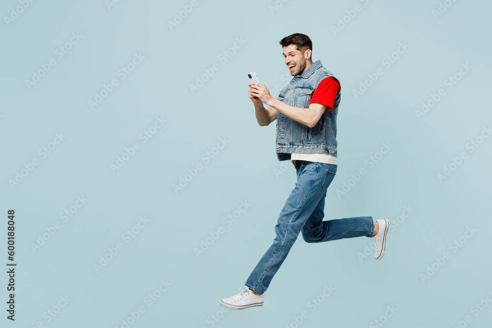 Full body side view young man he wears denim vest red t-shirt casual clothes jump high hold in hand use mobile cell phone isolated on plain pastel light blue cyan background studio. Lifestyle concept.
