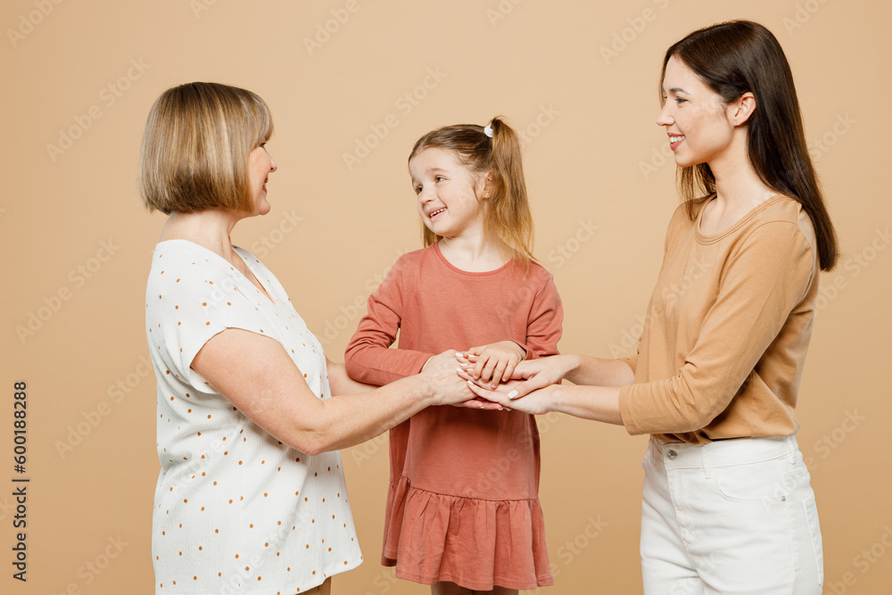 Happy smiling fun women wears casual clothes with child kid girl 6-7 years old. Granny mother daughter hold hands looking to each other isolated on plain beige background. Family parent day concept. Happy smiling fun women wears casual clothes with child kid girl 6-7 years old. Granny mother daughter hold hands looking to each other isolated on plain beige background. Family parent day concept.