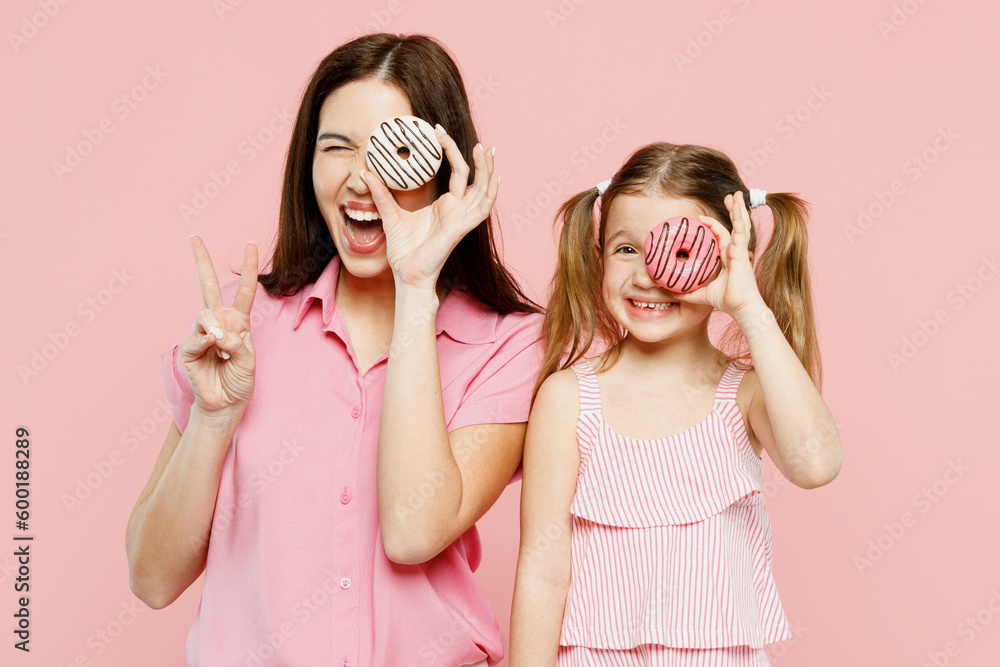 Happy smiling woman wear casual clothes with child kid girl 6-7 years old. Mother daughter hold cover eye with donuts, show v-sign isolated on plain pastel pink background. Family parent day concept. Happy smiling woman wear casual clothes with child kid girl 6-7 years old. Mother daughter hold cover eye with donuts, show v-sign isolated on plain pastel pink background. Family parent day concept.