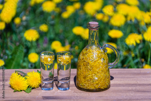 Homemade dandelion flowers tincture in two glasses and in a glass bottle on a wooden table in a summer garden, closeup