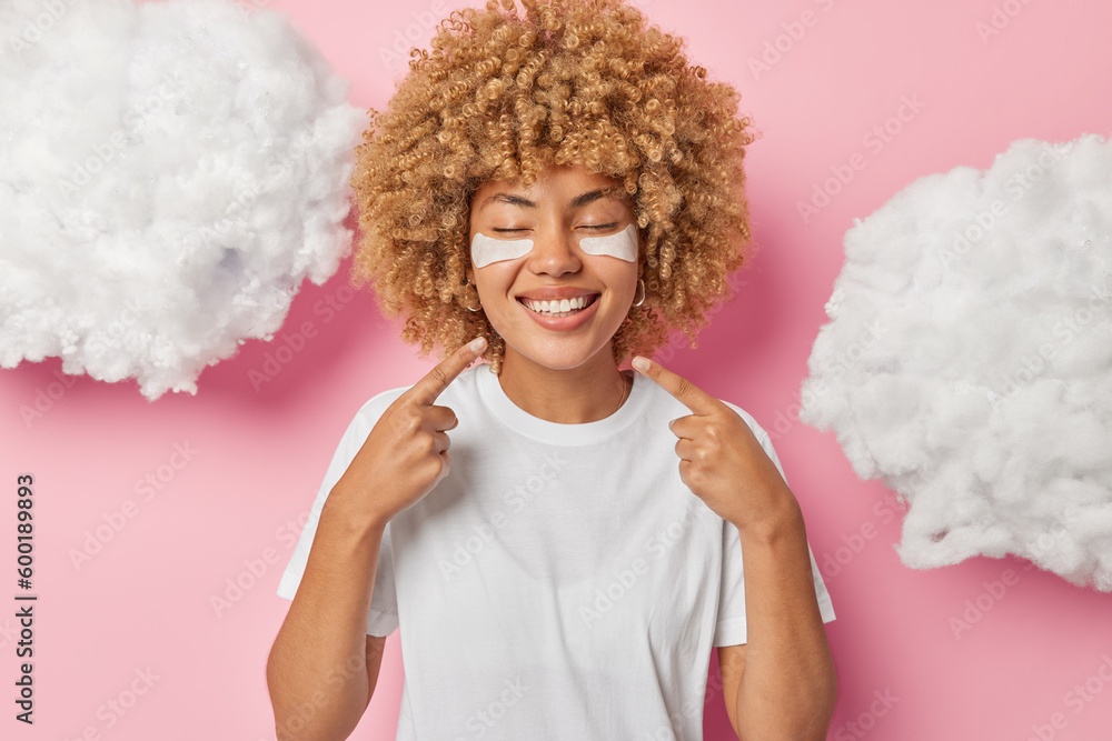 Look at my perfect smile. Cheerful young millennial woman smiles toothily points at her even teeth keeps eyes closed wears white t shirt undergoes skin care procedures isolated over pink background Look at my perfect smile. Cheerful young millennial woman smiles toothily points at her even teeth keeps eyes closed wears white t shirt undergoes skin care procedures isolated over pink background