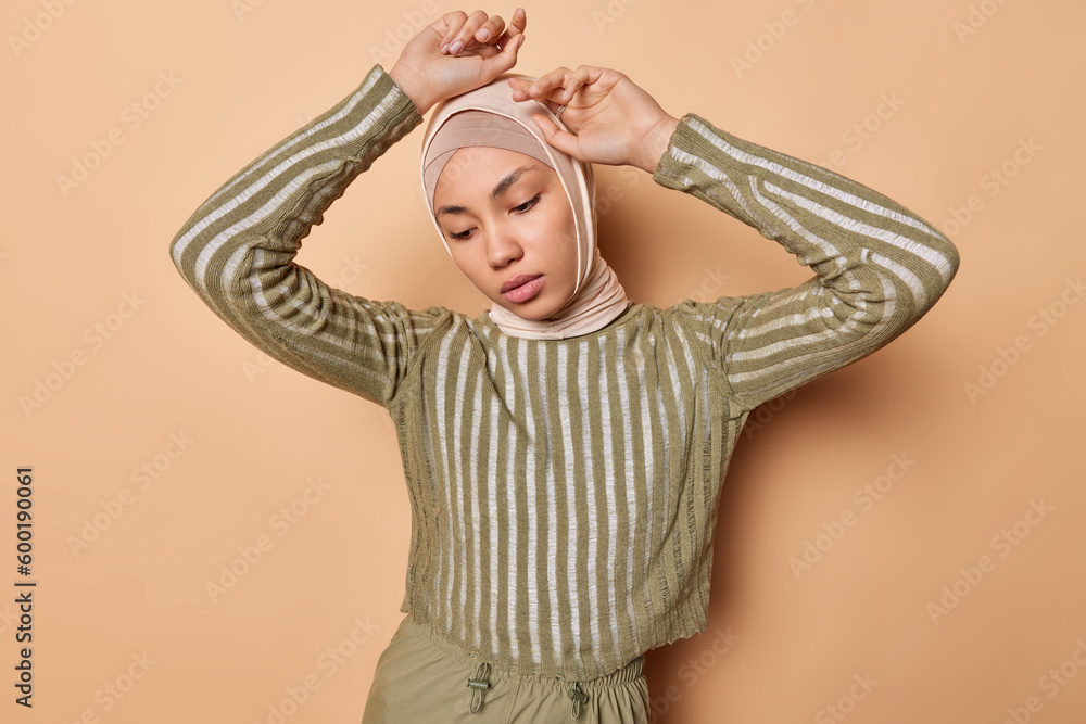 Φωτογραφία Thoughtful young Arabian woman keeps arms raised up over head concentrated wrapped in veil striped jumper concentrated down isolated over brown background