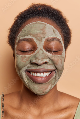 Beauty procedures concept. Close up shot of cheerful Afro American woman keeps eyes closed being in good mood smiles gently applies facial mud mask to reduce fine lines isolated over brown background