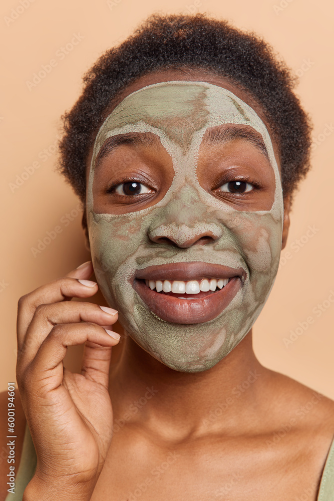 Fototapeta premium Headshot of young Afro American woman with short hair applies facial beauty mask for skin treatment smiles gently being in good mood focused directly at camera isolated over brown background.