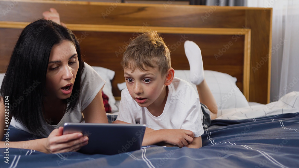 Front view of pretty loving young dark-haired mother which teaching her handsome joyful small son how to use tablet pc,lying on bed Front view of pretty loving young dark-haired mother which teaching her handsome joyful small son how to use tablet pc,lying on bed