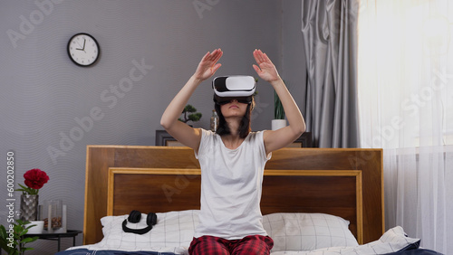 Close up of likable concentrated 30-aged woman in pajamas which sitting on comfortable bed in bedroom and making scrolling gestures on virtual screen applying augmented reality goggles photo