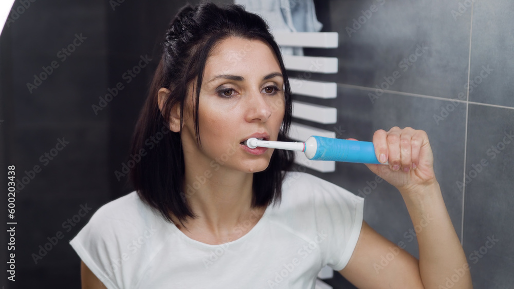 Front view of appealing satisfied young woman in t-shirt which looking at her reflection in the bathroom mirror during cleaning teeth Front view of appealing satisfied young woman in t-shirt which looking at her reflection in the bathroom mirror during cleaning teeth