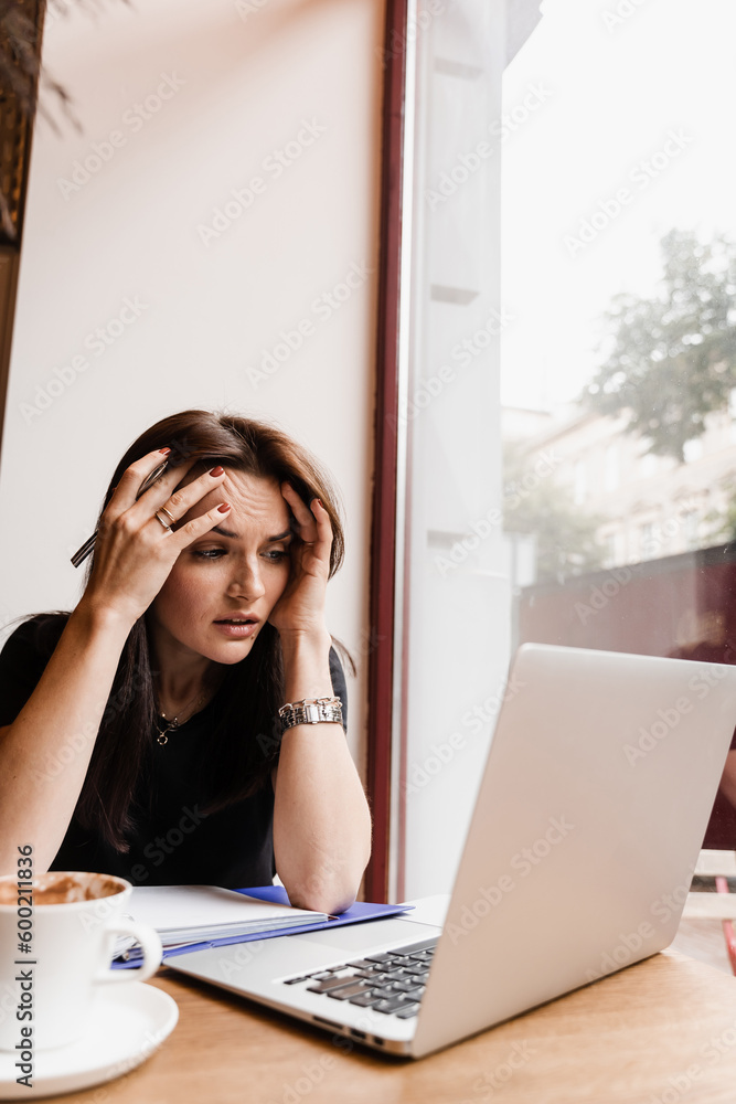Woman manager with strong headache working on laptop online remotely in loft cafe. Sad girl with laptop got migraine touches her head because of pain.