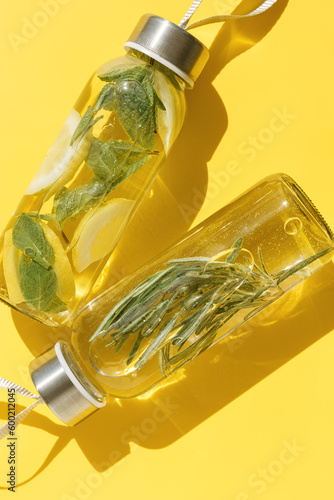 Top view closeup two glass bottles, water drink detox with lemon and mint leaves, and rosemary, at sunlight on yellow color background. Infused water, wellness trend. Refreshing summer drink