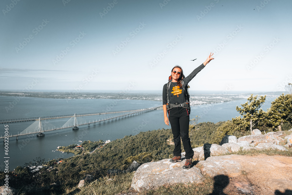 Obraz premium Mulher mochileira posando para foto no alto da montanha com paisagem ao fundo. Imagem com espaço para texto.