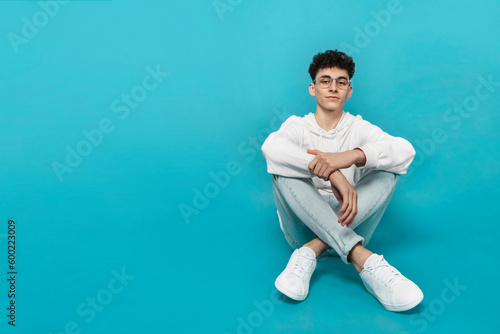 Foto Confident teen boy in eyewear and white clothes, sitting down, looking at camera, isolated  turquoise background