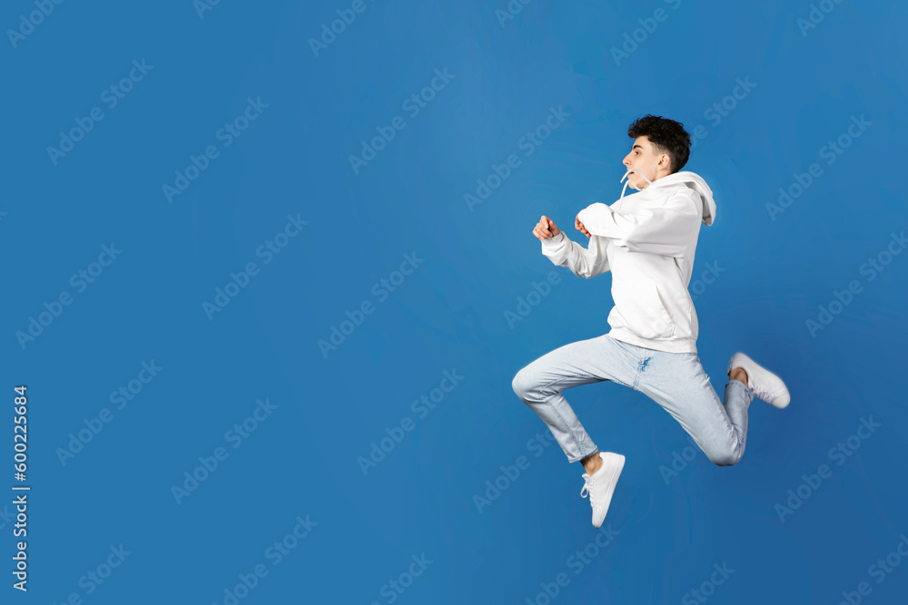 Flull length profile image of a jumping teen boy in white hoodie and jeans, isolated blue background. Space for text.