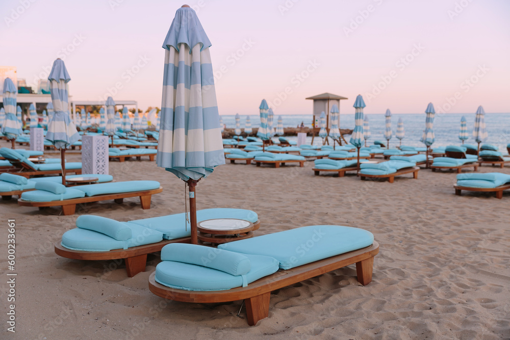 Evening beach at sunset. Empty blue wooden sunbeds and umbrellas on a sandy beach near the sea