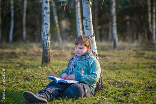 A handsome boy in a scarf is in a spring park, sitting under a birch tree, reading a book and enjoying his dreams. © Alina Lebed