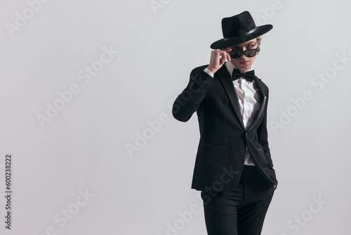 elegant young groom in black tuxedo wearing hat and adjusting sunglasses