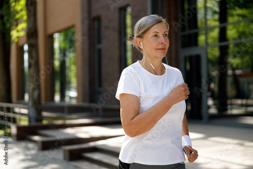 Side view of the smiled Mature Woman running and wearing headphones in the street. Sport and healthy lifestyle concept