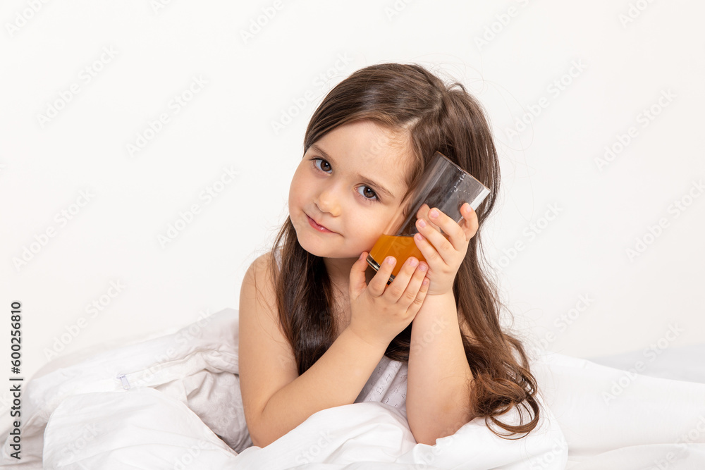 Happy little girl child in pajamas with a glass of fresh orange juice sitting at home in bed. Good morning Happy little girl child in pajamas with a glass of fresh orange juice sitting at home in bed. Good morning