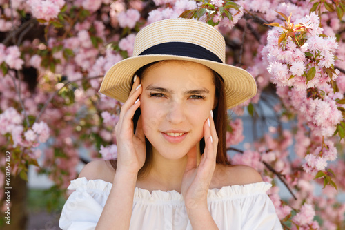 Close up Portrait of the Smiling Brunette Girl Posing, Touching Her Face on the Sakura Tree Blooming Outside. Femininity and Natural Beauty Concept