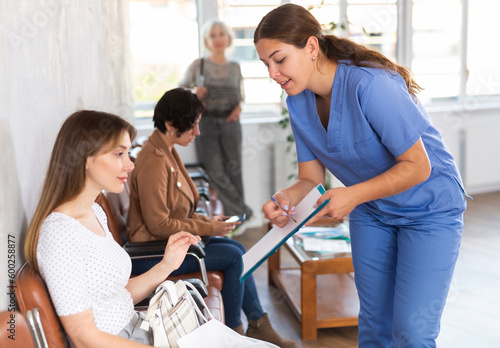 Friendly young woman  professional doctor wearing blue uniform  showing papers while explaining results of medical examination to female patient sitting in lobby of clinic