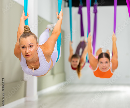 Focused sporty woman performing locust pose, balancing and stretching Salabhasana asana with arched back in hanging hammock during aerial yoga group training in studio photo