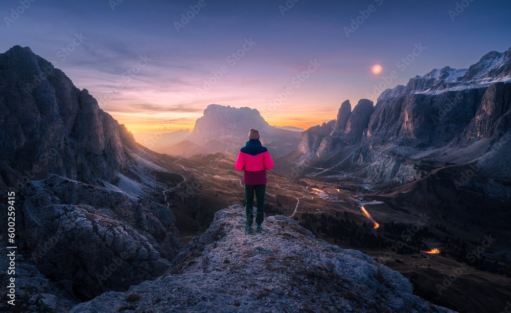 Woman on the rock and mountain peaks at night in autumn in Dolomites, Italy. Girl on the stone, high rocks, sky with moon, light trails on road at twilight in fall. Colorful landscape with cliffs
