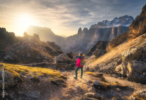 Girl with backpack on the trail in mountain canyon at sunset in autumn. Beautiful landscape with young woman, high rocks, path, stones, orange grass, sky in fall in Dolomites, Italy. Hiking. Nature