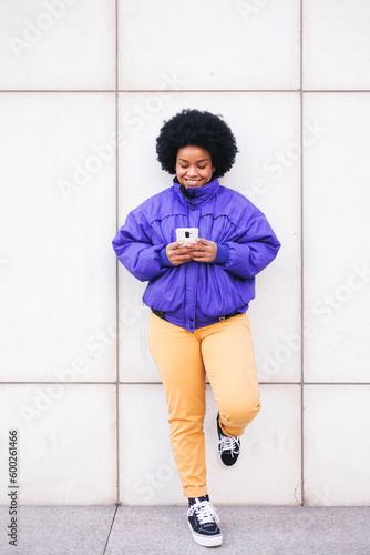 young latin afro woman standing on white wall using phone and smiling