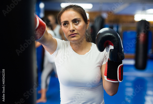 Latin woman in boxing gloves beating punchbag during group boxing training. © JackF