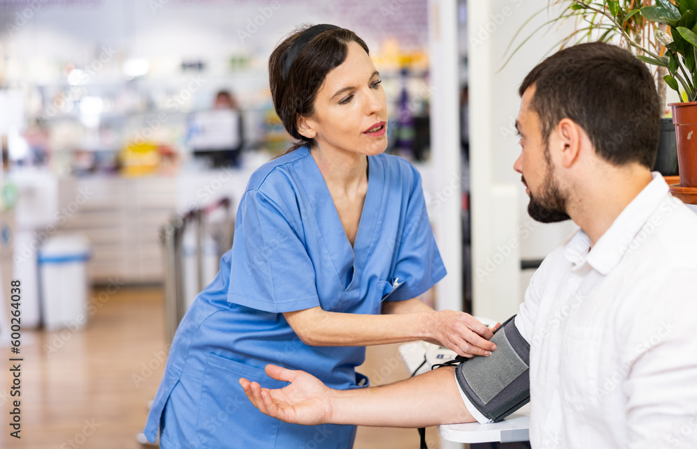 Obraz premium Portrait of positive female pharmacist checking blood pressure of male customer at pharmacy
