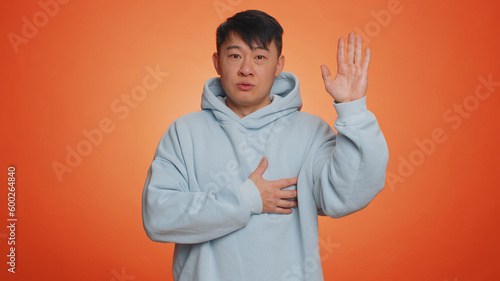 I swear to be honest. Sincere responsible asian handsome man raising hand to take oath, promising to be honest and to tell truth, keeping hand on chest. Adult guy isolated on orange studio background