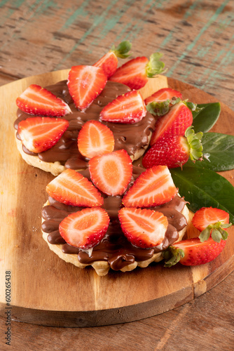 dessert of two waffles with chocolate and hazelnut cream on a round wooden board and cut strawberries on a wooden table.at a close-up angle