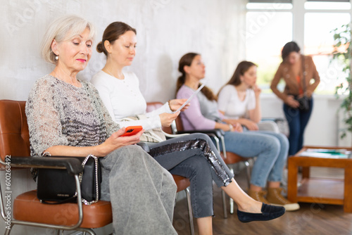 Positive senior woman browsing websites or chatting with friends on smartphone while sitting in lobby of public service building or medical facility, waiting for appointment