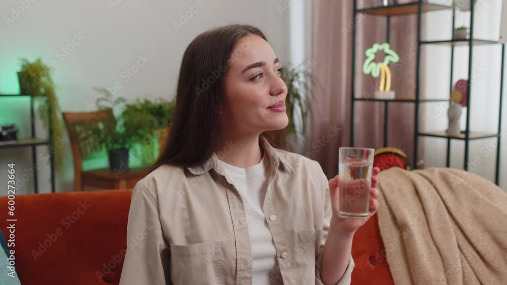 Thirsty young woman holding glass of natural aqua make sips drinking still water preventing dehydration sits at home living room. Girl with good life habits, healthy slimming, weight loss concept Thirsty young woman holding glass of natural aqua make sips drinking still water preventing dehydration sits at home living room. Girl with good life habits, healthy slimming, weight loss concept