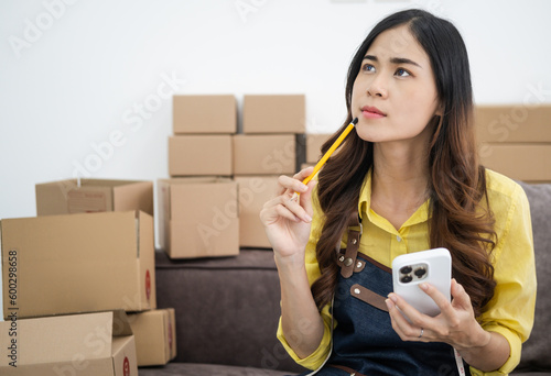 Young asian online seller businesswoman working shipping at home looking at mobile phone for online orders and thinking about her business future, good for online seller business concept.