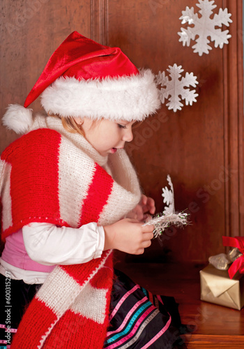 A girl in a warm red scarf and a santa claus hat examines paper snowflakes. The child rejoices in winter, New Year cartoons and the holiday of Christmas