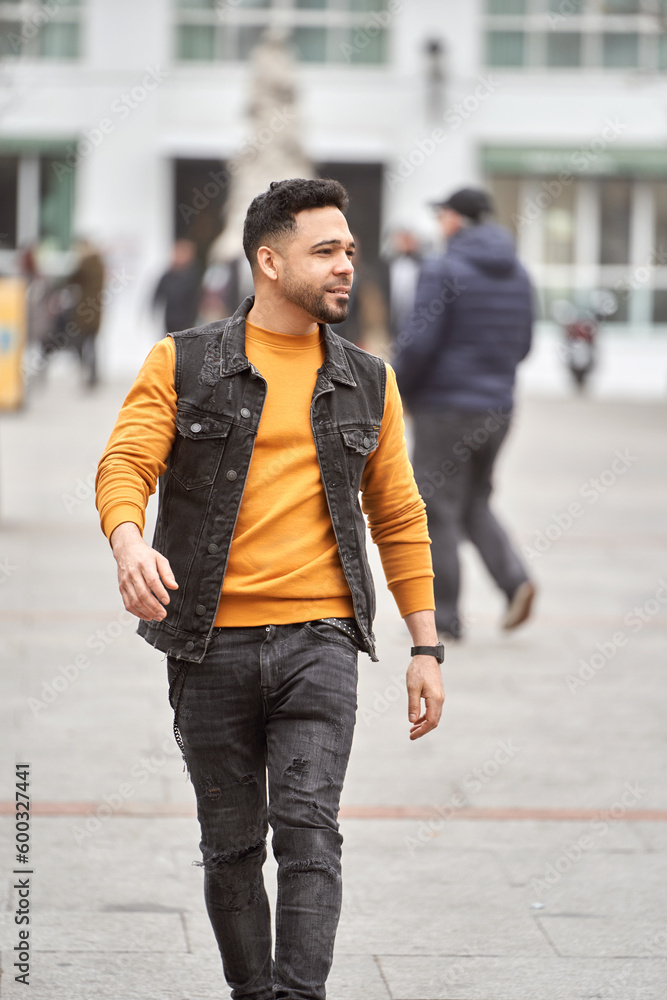 Latin man walking through the city streets wearing orange sweater and black vest. Latin man walking through the city streets wearing orange sweater and black vest.
