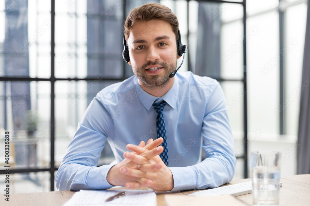 Cheerful young support phone male operator in headset, at workplace with collegues on the background while using computer, help service and client consulting call center concept. Cheerful young support phone male operator in headset, at workplace with collegues on the background while using computer, help service and client consulting call center concept.