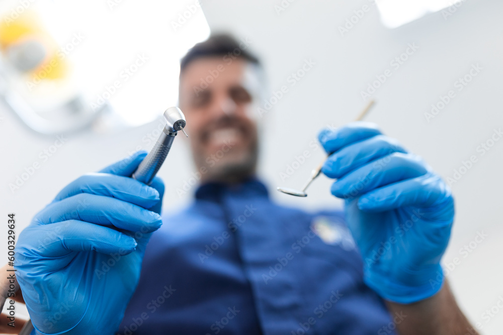 Dental instruments in the hands of a blurred male dentist doing dental examination. Dentist holding angled mirror and dental drill bending over patient Dental instruments in the hands of a blurred male dentist doing dental examination. Dentist holding angled mirror and dental drill bending over patient