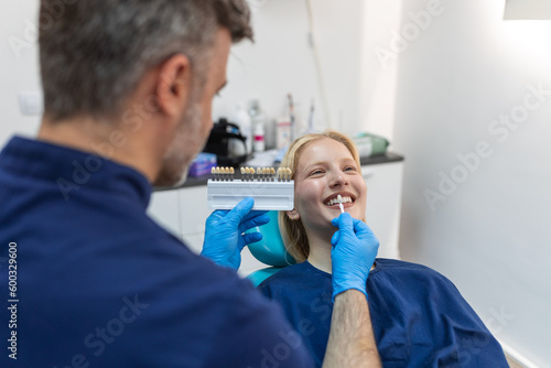 Image of satisfied young woman sitting in dental chair at medical center while professional doctor fixing her teeth  Female dentist choosing filling shade for smiling woman  using tooth scale sample