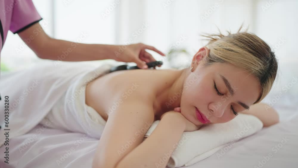 Wellness body massage and spa concept. Woman masseur putting hot stone on back of woman in white towel lying on spa bed for treatment therapy at spa salon Wellness body massage and spa concept. Woman masseur putting hot stone on back of woman in white towel lying on spa bed for treatment therapy at spa salon