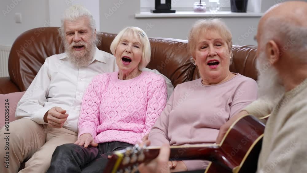 Group of relaxed senior friends singing as talented bearded man playing guitar. Happy positive Caucasian retirees having fun meeting indoors