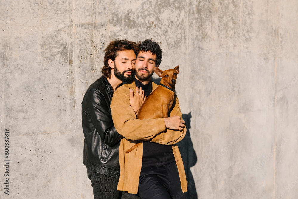 Gay couple with eyes closed carrying dog in front of concrete wall Gay couple with eyes closed carrying dog in front of concrete wall
