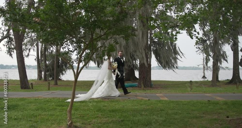 Side view of bride and groom walking on side walk with the lake behind them in slow motion while looking at each other photo