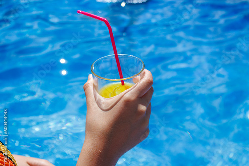  Unrecognizable person with transparent glass of fresh cold orange juice with ice and red straw against blue clear water of swimming pool in sunny resort 