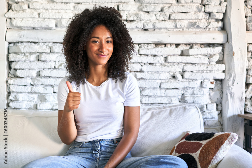 A young african american woman sitting on her sofa smiling and raising thumb up A young african american woman sitting on her sofa smiling and raising thumb up