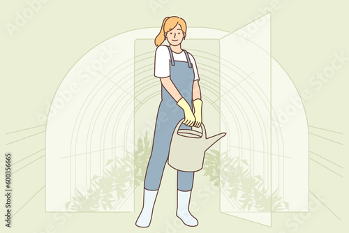 Woman gardener comes out of greenhouse made of glass and holds watering can filled with fertilizer