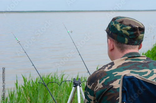 A young man is fishing. A man in camouflage clothes sits on the shore next to fishing rods.