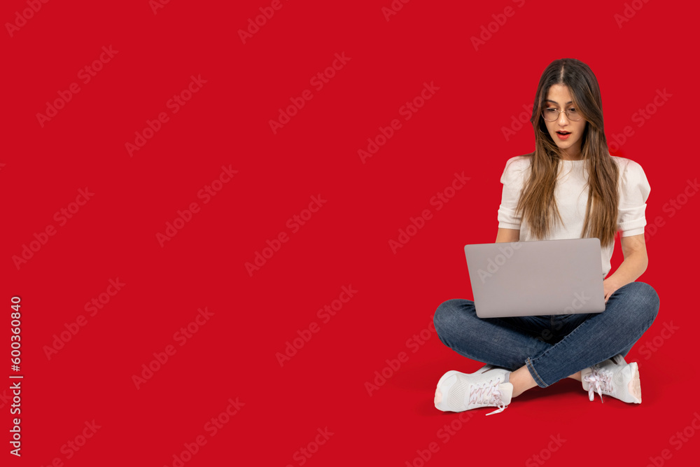 Shocked woman, full body length view of shocked woman. Brunette young girl sitting on the ground. Holding laptop looking screen surprised what she see. Isolated red background. Copy space. 